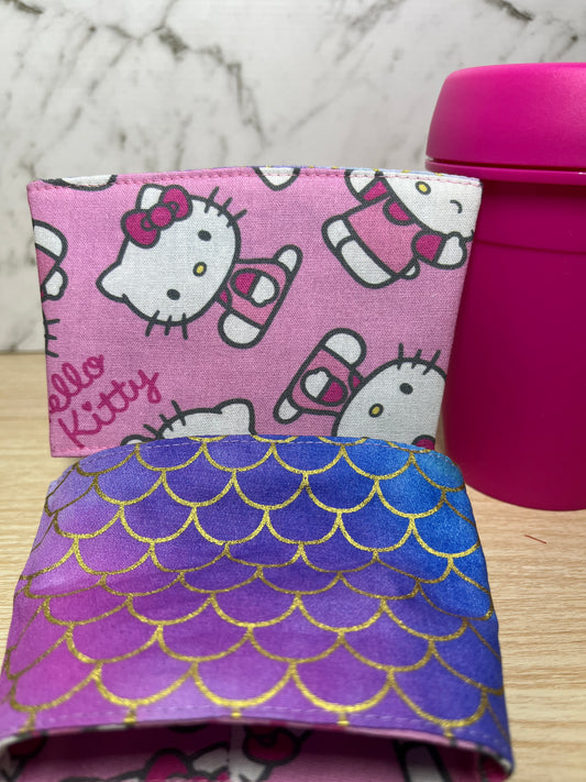 Two fabric cup sleeves, one with Hello Kitty pattern and the other with mermaid scale design, on a marble surface.