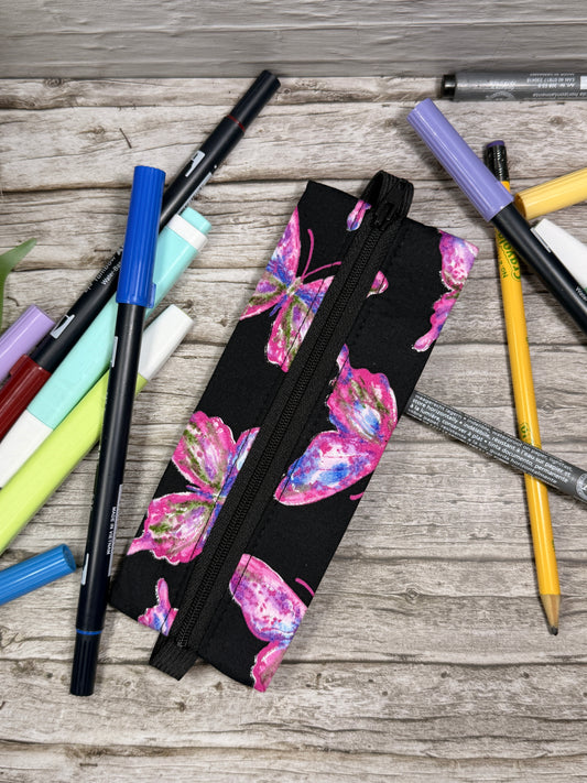 Pencil case with butterfly design on a notebook which is on a wooden surface with pens and markers.