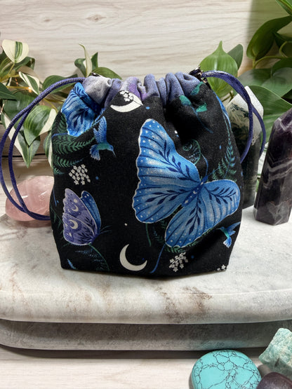Black drawstring bag with blue butterfly designs on a light wooden surface with greenery.