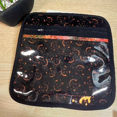 Black cross stitch project bag with orange cat faces patterns on a wooden surface next to a plant