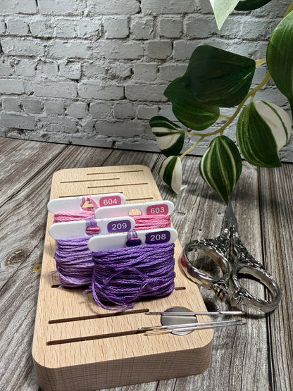 Purple and pink floss on a wooden floss board with needles, scissors and a plant in the background