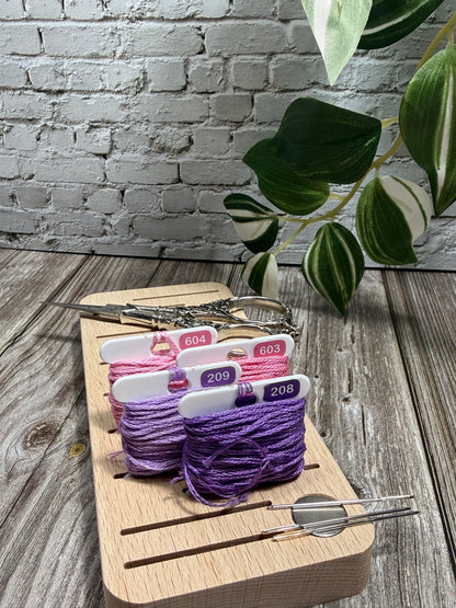 Purple and pink floss on a wooden floss board with needles, scissors and a plant in the background