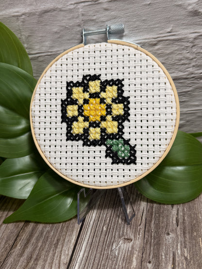 A circular embroidery hoop holding white cross-stitch aida fabric with a floral design stitched in black, yellow, and green, displayed on green leaves over a rustic wood surface.