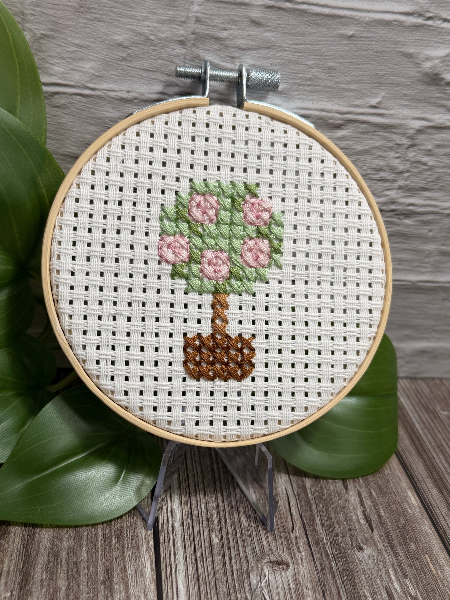 A completed cross-stitch embroidery in a wooden hoop, featuring a small potted plant with pink flowers and green leaves on white Aida fabric, displayed on a stand with green leaves and a gray brick wall in the background.