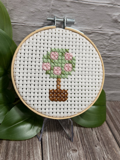 A completed cross-stitch embroidery in a wooden hoop, featuring a small potted plant with pink flowers and green leaves on white Aida fabric, displayed on a stand with green leaves and a gray brick wall in the background.