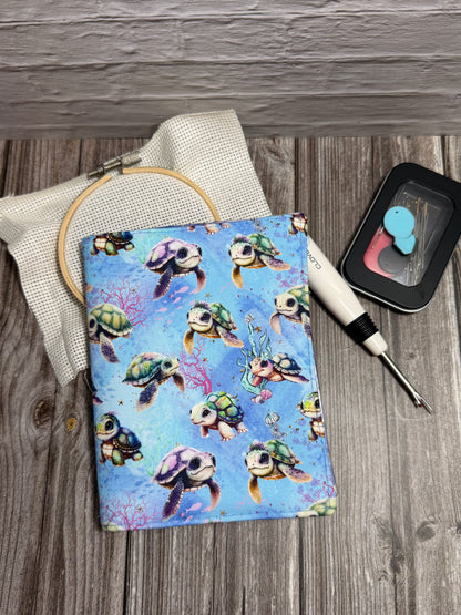 A needle book with a colorful turtle print on a blue background, styled on a gray wood-grain surface with a seam ripper, embroidery hoop, and small black needle case.