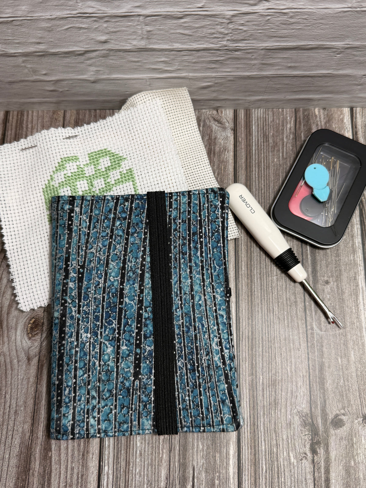 A fabric needle book with embroidery tools on a wood surface, holding scissors, a white-handled seam ripper, and a white embroidery canvas with green stitching. A black needle case is partially visible behind the needle book.