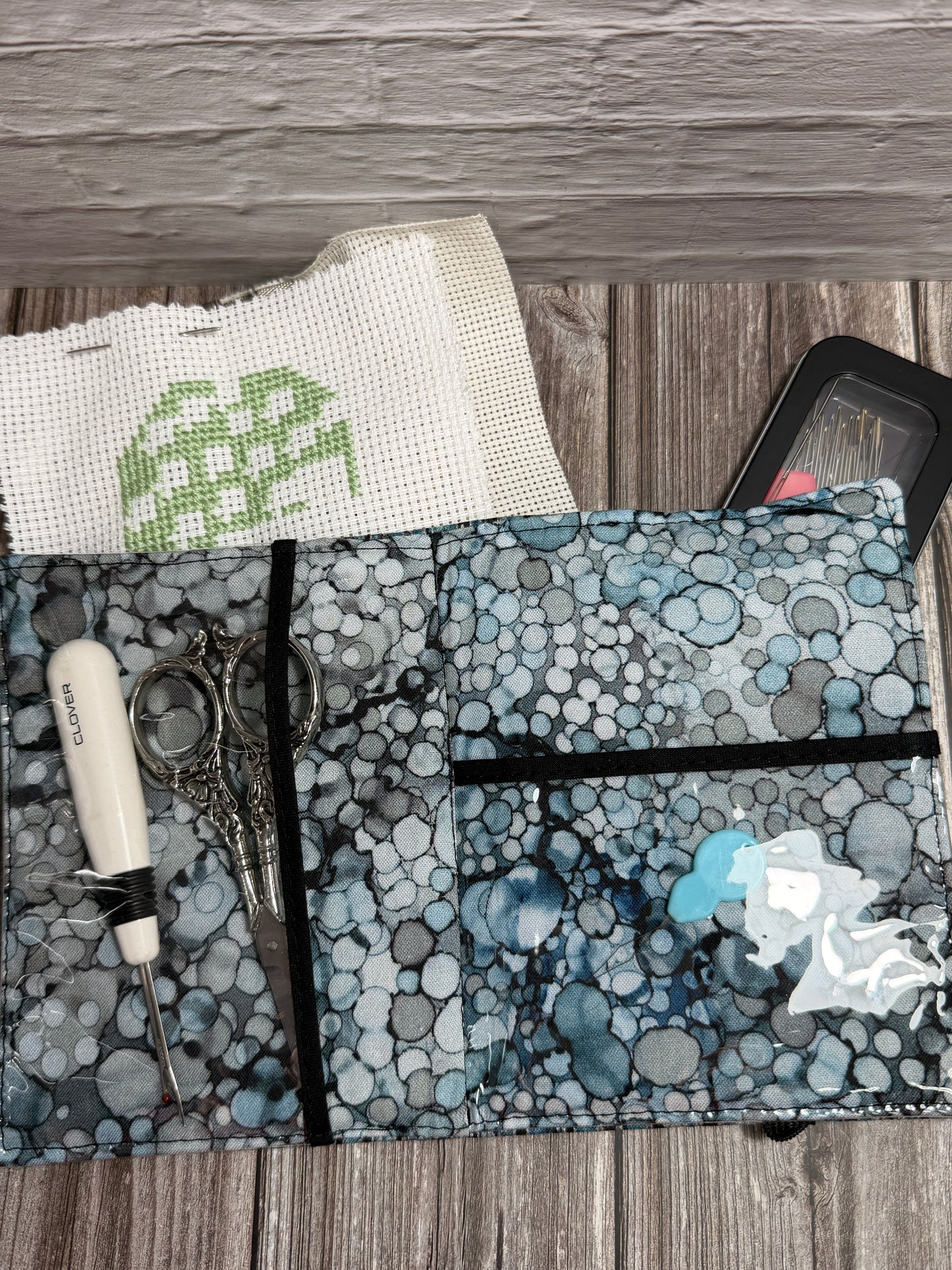 A fabric needle book with embroidery tools on a wood surface, holding scissors, a white-handled seam ripper, and a white embroidery canvas with green stitching. A black needle case is partially visible behind the needle book.