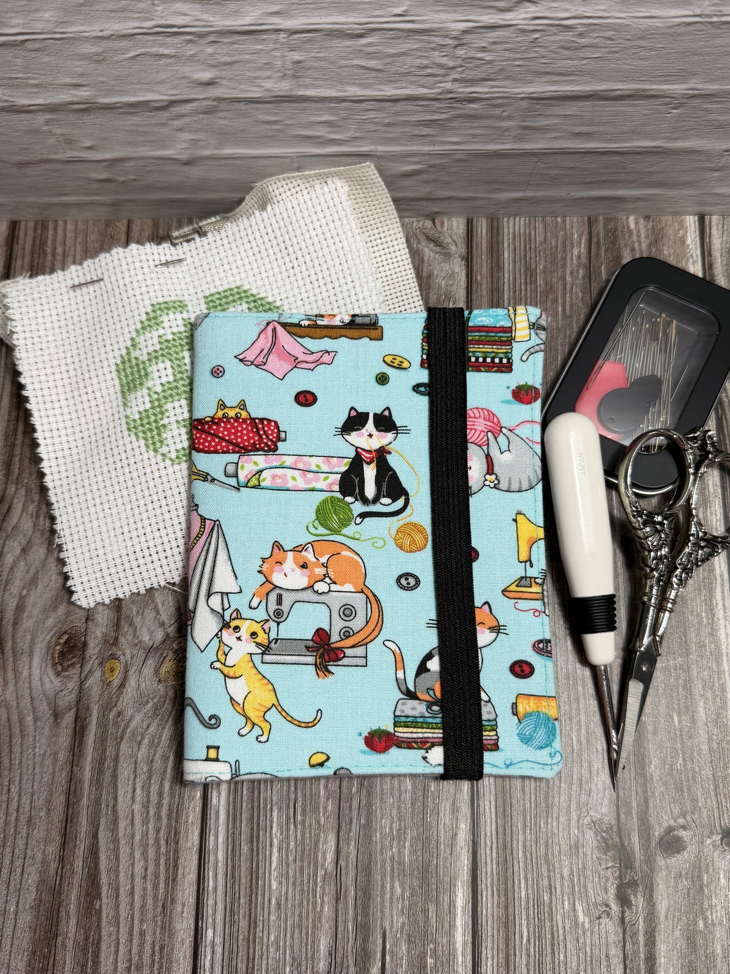 A needle book with a light blue background and a colorful cartoon cat sewing pattern print, secured with a black elastic band. It is styled on a gray wood background with a white aida fabric, scissors, and a small black needle case.