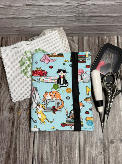 A needle book with a light blue background and a colorful cartoon cat sewing pattern print, secured with a black elastic band. It is styled on a gray wood background with a white aida fabric, scissors, and a small black needle case.