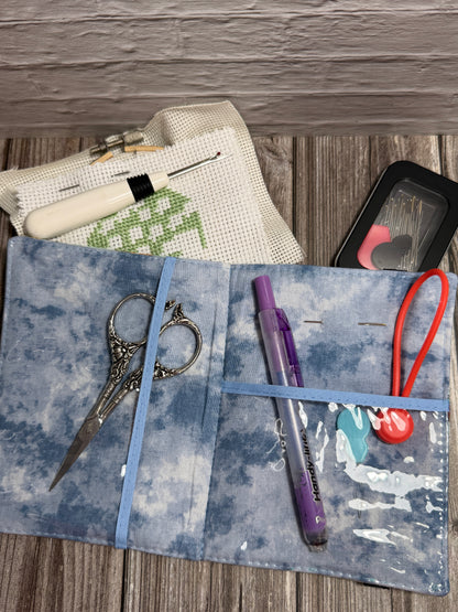 A fabric needle book with embroidery tools on a wood surface, holding scissors, a white-handled seam ripper, and a white embroidery canvas with green stitching. A black needle case is partially visible behind the needle book.