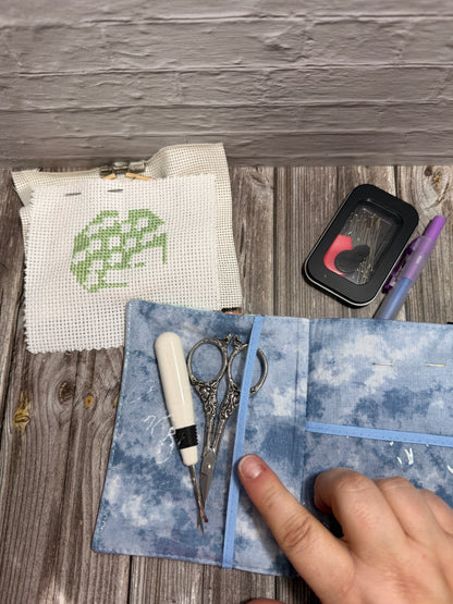 A close-up of a blue fabric needle case with elastic loops holding embroidery scissors and a white-handled needle, with a person's hand pointing at it. A small embroidered fabric sample and a black case with a purple pen are arranged on a rustic wood surface.