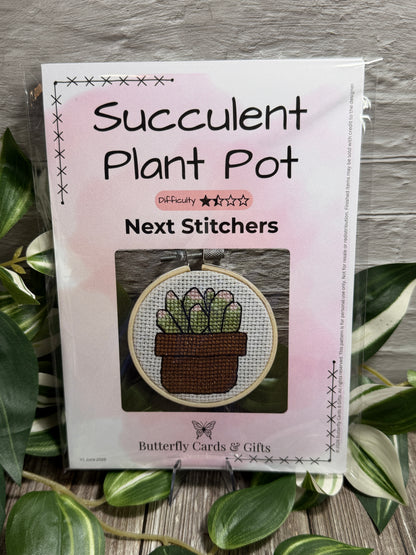 A packaged cross-stitch pattern titled 'Succulent Plant Pot' from Butterfly Cards and Gifts, displayed on a rustic wood surface surrounded by green plant leaves. The package is white with a pink watercolor wash and shows a sample stitched design of a succulent in a pot.