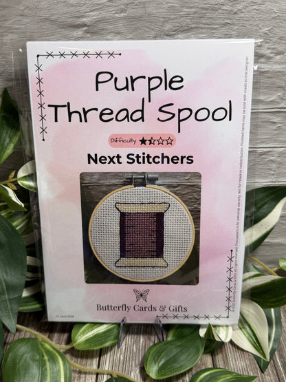 A cross stitch kit titled 'Purple Thread Spool' from Next Stitchers range, showing a small embroidery hoop with purple thread, photographed on a wood surface with green leaves around it.