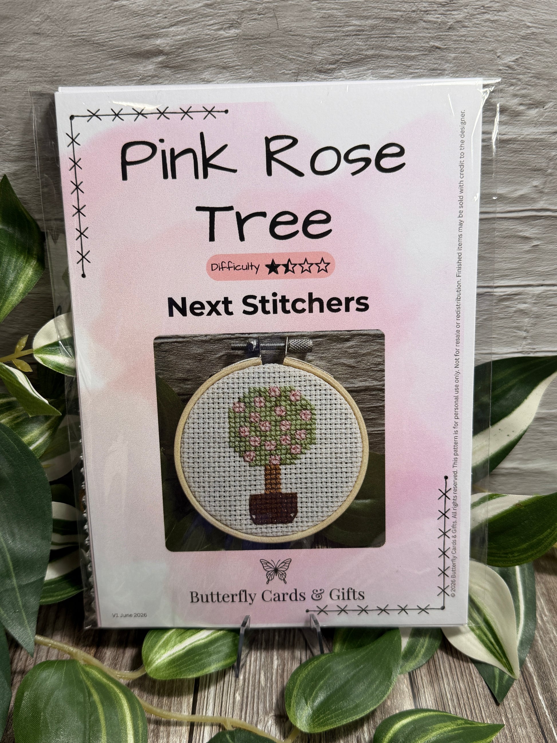 A packaged cross stitch kit titled 'Pink Rose Tree Next Stitches' by Butterfly Cards & Gifts, shown in a clear plastic sleeve on a gray wood surface surrounded by green variegated leaves. The package features a pink watercolor design and a photo of a completed cross-stitch tree in a hoop.