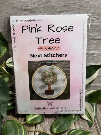 A packaged cross stitch kit titled 'Pink Rose Tree Next Stitches' by Butterfly Cards & Gifts, shown in a clear plastic sleeve on a gray wood surface surrounded by green variegated leaves. The package features a pink watercolor design and a photo of a completed cross-stitch tree in a hoop.