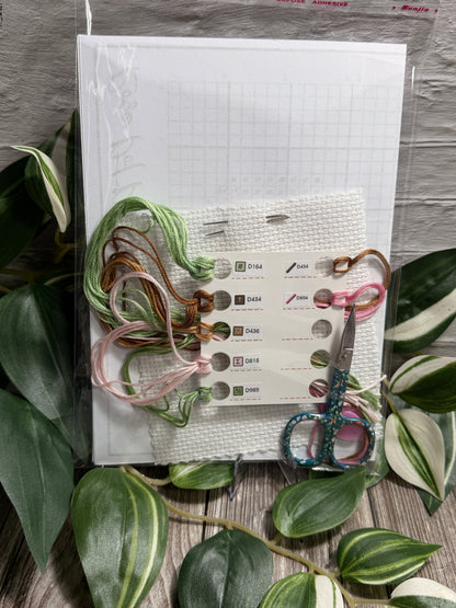 A flat-lay of a cross stitch kit featuring a white grid paper pattern, aida fabric, needles a white mesh card with color swatches, several strands of embroidery floss in green, pink, and brown, and a pair of patterned scissors, all arranged on a gray wood-textured surface with green leaves.