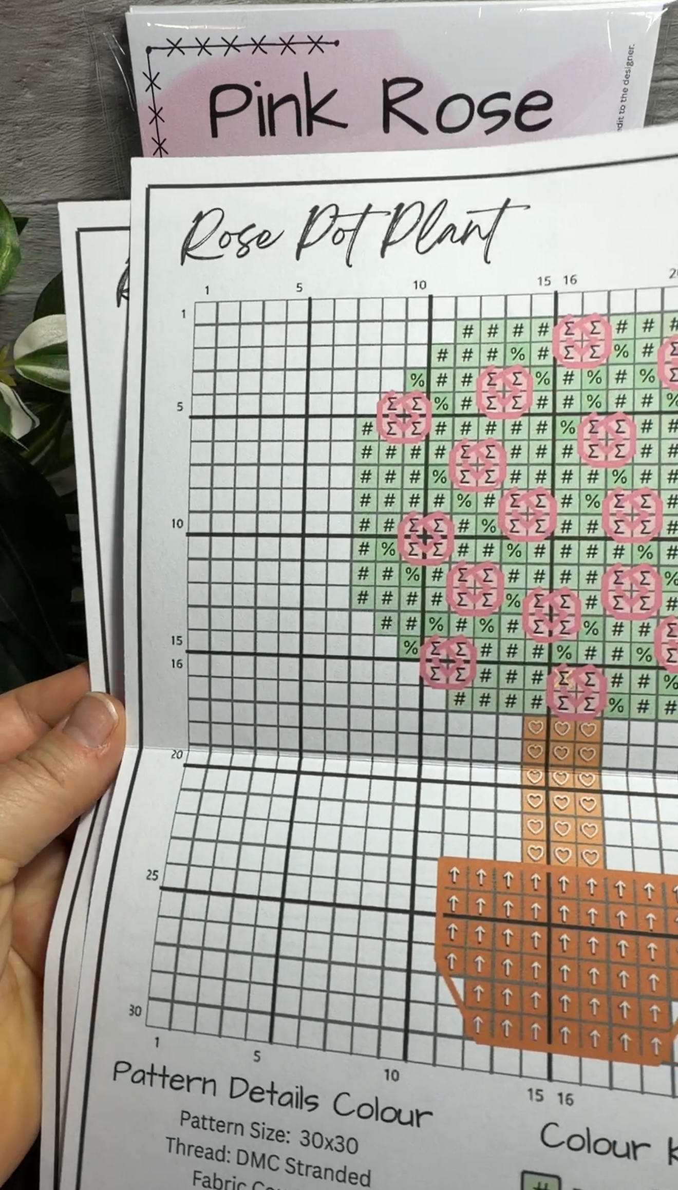 A close-up of a hand holding a printed cross stitch pattern sheet titled 'Pink Rose Rose Pot Plant,' showing colored fabric squares arranged to form a rose and pot design.