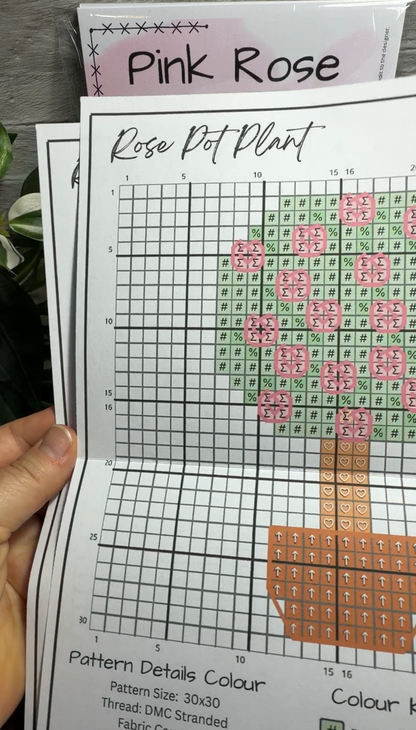 A close-up of a hand holding a printed cross stitch pattern sheet titled 'Pink Rose Rose Pot Plant,' showing colored fabric squares arranged to form a rose and pot design.