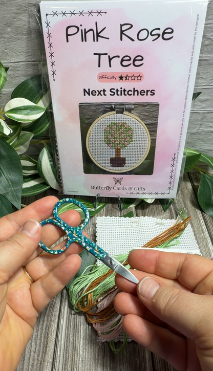 A person holds teal-handled embroidery scissors in front of a pink 'Pink Rose Tree Next Stitches' kit box, with thread and a canvas visible on a wood surface.