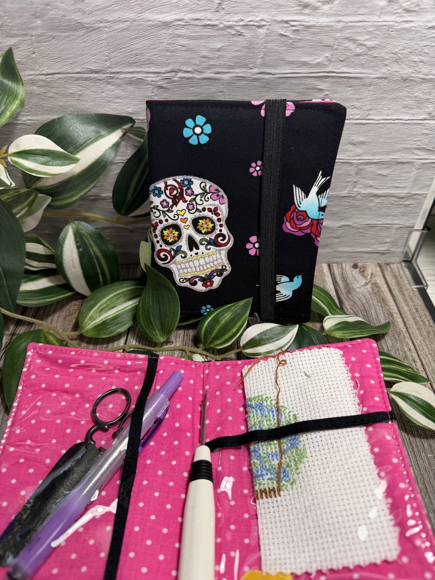 A fabric needle book (needlework) kit shown open and closed on a rustic wood surface with variegated green leaves. The closed view shows a black fabric cover with a colorful sugar skull and floral print; the open view shows a bright pink fabric interior with white polka dots, a white cross-stitch canvas, thread, and tools including scissors, a pen, a seam ripper and a needle threader.