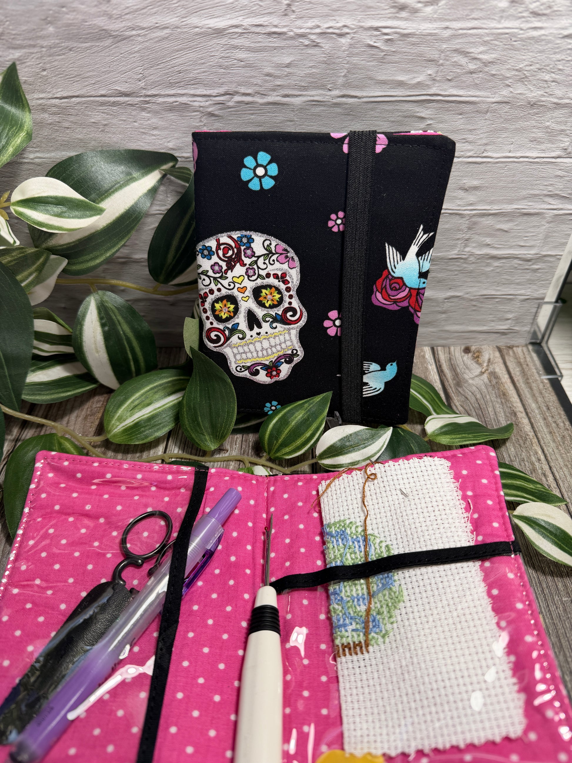 A fabric needle book (needlework) kit shown open and closed on a rustic wood surface with variegated green leaves. The closed view shows a black fabric cover with a colorful sugar skull and floral print; the open view shows a bright pink fabric interior with white polka dots, a white cross-stitch canvas, thread, and tools including scissors, a pen, a seam ripper and a needle threader.