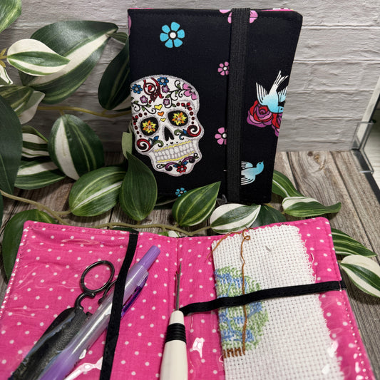 A fabric needle book (needlework) kit shown open and closed on a rustic wood surface with variegated green leaves. The closed view shows a black fabric cover with a colorful sugar skull and floral print; the open view shows a bright pink fabric interior with white polka dots, a white cross-stitch canvas, thread, and tools including scissors, a pen, a seam ripper and a needle threader.