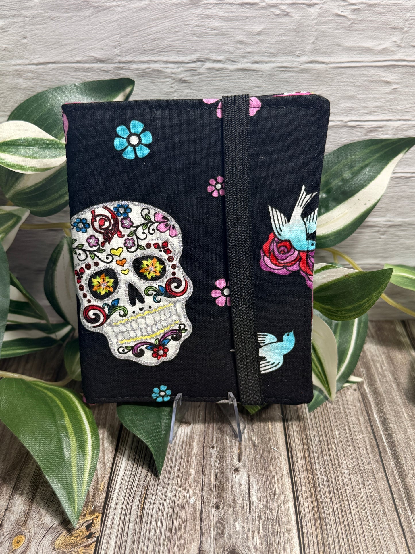 A needle book with a colorful sugar skull on a black fabric, secured with a black elastic band, displayed on a wooden surface with green variegated leaves and a seam ripper and embroidery scissors near by.