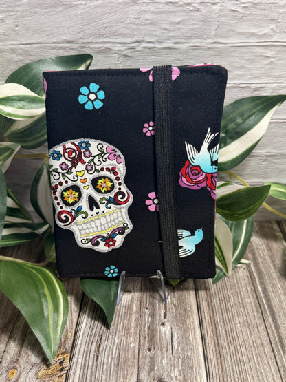 A needle book with a colorful sugar skull on a black fabric, secured with a black elastic band, displayed on a wooden surface with green variegated leaves and a seam ripper and embroidery scissors near by.