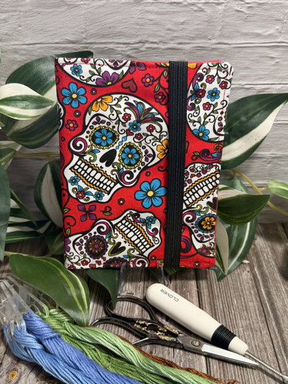 A needle book with a colorful sugar skull and floral Day of the Dead pattern on a red background, shown standing upright with a black elastic band closure. The case is styled on a wooden surface with green leaves, embroidery floss, seam ripper and scissors.