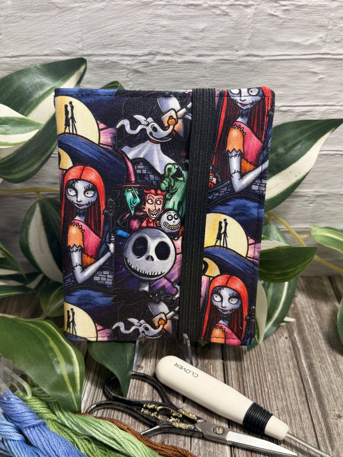 A needle book featuring a colorful 'The Nightmare Before Christmas' character print, displayed on a rustic wood surface with green leaves, embroidery floss, and scissors.