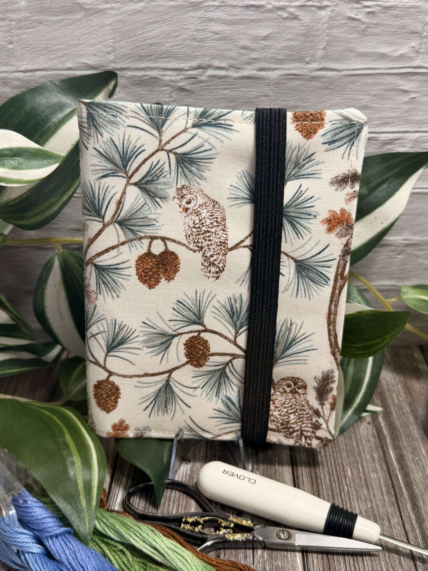 A needle book with an owl and pine tree print, standing upright on a rustic wooden surface with embroidery floss, seam ripper and embroidery scissors.