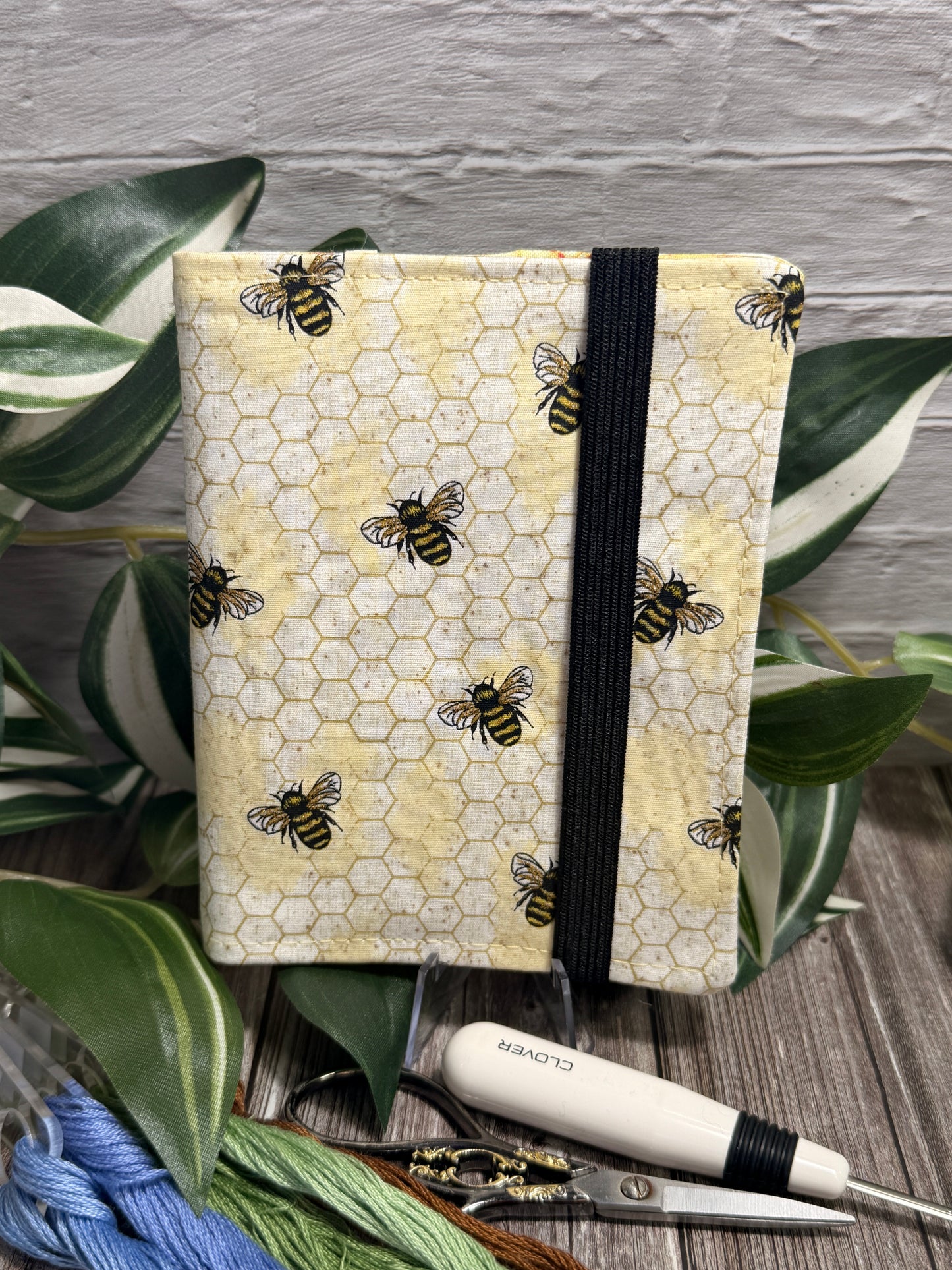 A needle book with a yellow honeycomb pattern and printed bees, bound with a black elastic band. It is styled on a wooden surface with green leaves, embroidery floss, and a white-handled seam ripper and embroidery scissors.
