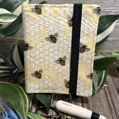 A needle book with a yellow honeycomb pattern and printed bees, bound with a black elastic band. It is styled on a wooden surface with green leaves, embroidery floss, and a white-handled seam ripper and embroidery scissors.