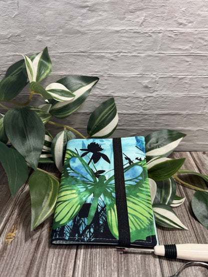 A needle book with a Butterfly and leaf print in green, blue, and black, secured with a black elastic band, displayed on a wooden surface with green variegated leaves and a seam ripper and embroidery scissors near by.