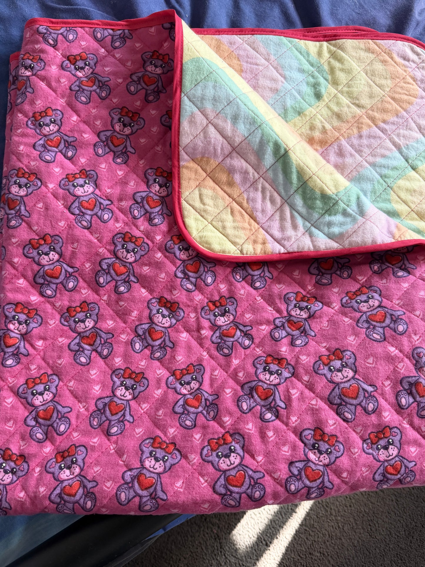 A folded quilted blanket or comforter with a pink front side featuring a repeating teddy bear pattern with red hearts, and a pastel multicolor back side with a dark pink border.