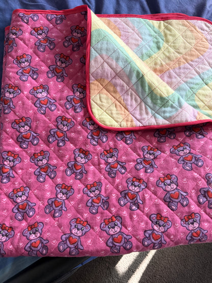 A folded quilted blanket or comforter with a pink front side featuring a repeating teddy bear pattern with red hearts, and a pastel multicolor back side with a dark pink border.
