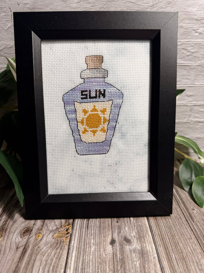 A framed cross stitched artwork depicting a bottle labeled 'SUN' with a yellow sun motif on a white aida  background, set in a black frame on a wooden surface with green leaves and a brick wall behind it.