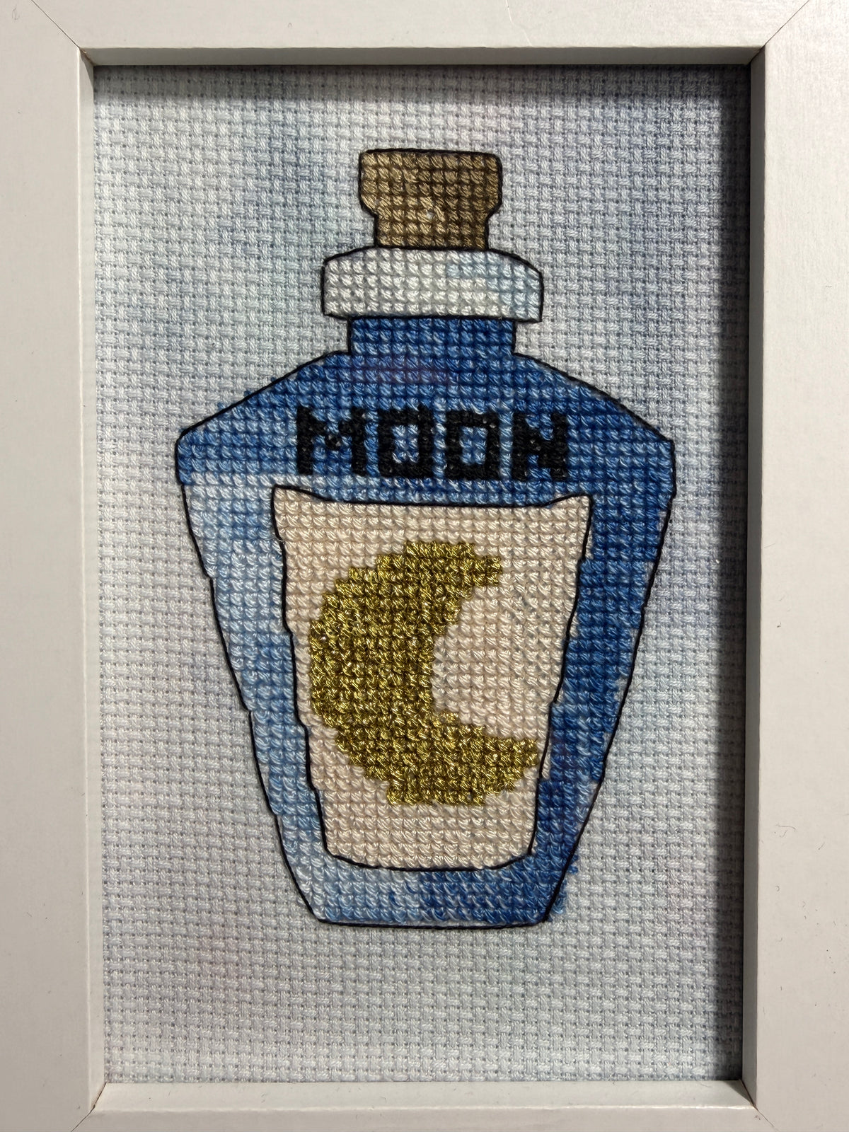 A framed cross-stitch artwork depicting a blue bottle labeled 'MOON' with a gold crescent moon on the label, set against a hand dyed aida fabric background.