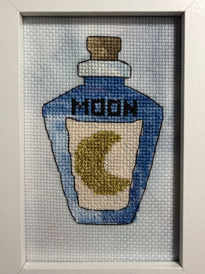 A framed cross-stitch artwork depicting a blue bottle labeled 'MOON' with a gold crescent moon on the label, set against a hand dyed aida fabric background.