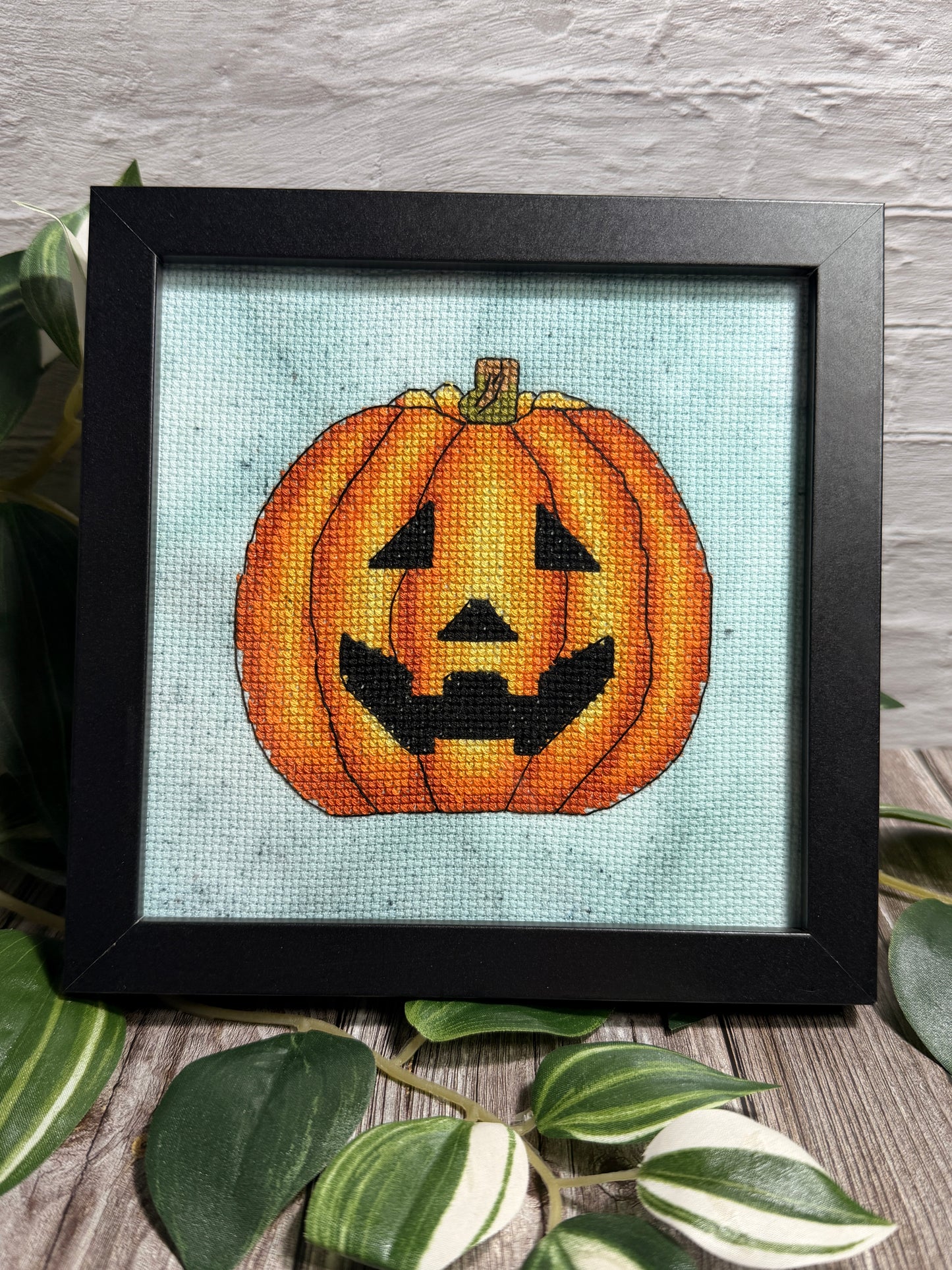A framed cross-stitch artwork featuring a carved orange pumpkin face on a hand dyed aida fabric background, set in a black frame and styled with green leaves on a wooden surface against a white brick wall.