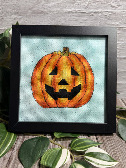 A framed cross-stitch artwork featuring a carved orange pumpkin face on a hand dyed aida fabric background, set in a black frame and styled with green leaves on a wooden surface against a white brick wall.