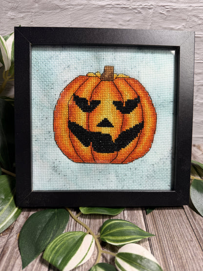 A framed cross-stitch artwork featuring a carved orange jack-o'-lantern pumpkin that has fangs on a hand dyed pale teal aida fabric background, displayed in a black frame on a wooden surface with green leaves around it.