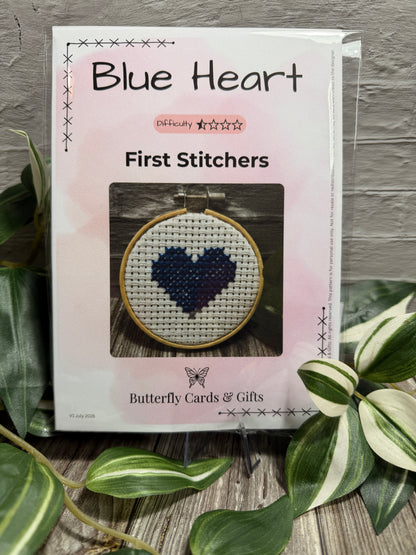 A packaged cross-stitch pattern titled "Blue Heart" by Butterfly Cards & Gifts, shown in a clear plastic sleeve on a tabletop with decorative green leaves around it. The front of the package features a photo of a completed circular cross-stitch design with a blue heart on white aida fabric.