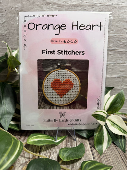 Packaged 'Orange Heart First Stitches' kit by Butterfly Cards & Gifts, displayed on a small stand with green variegated plant leaves in the foreground against a light gray brick wall.