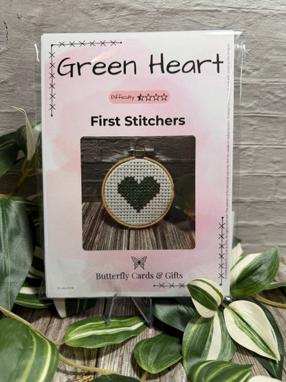 Packaged cross-stitch pattern kit titled 'Green Heart' in the First Stitches range, displayed on a small stand with decorative green leaves in the foreground against a gray brick wall background.