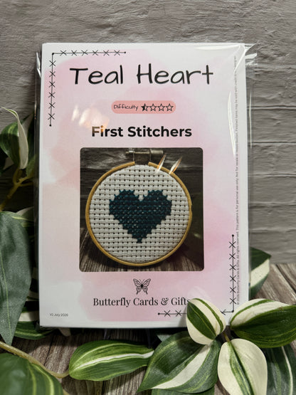 A packaged cross-stitch kit labeled 'Teal Heart' in the first stitchers range, shown in its clear plastic sleeve with a pink cover. The front displays a sample stitched heart design inside an embroidery hoop.