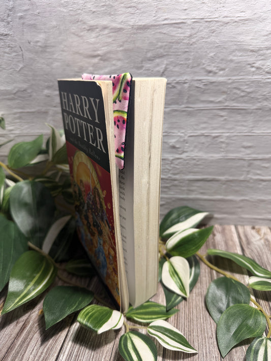 A Harry Potter book standing upright on a wooden surface, surrounded by green variegated houseplant leaves against a light gray brick wall. The book spine and cover are visible, with a colorful patterned bookmark peeking out from the top.