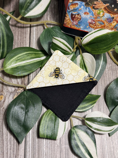 A square fabric book mark laid on a wood surface, surrounded by green variegated leaves. The top flap has a pale yellow honeycomb pattern with bee illustrations, and the lower section is solid black.