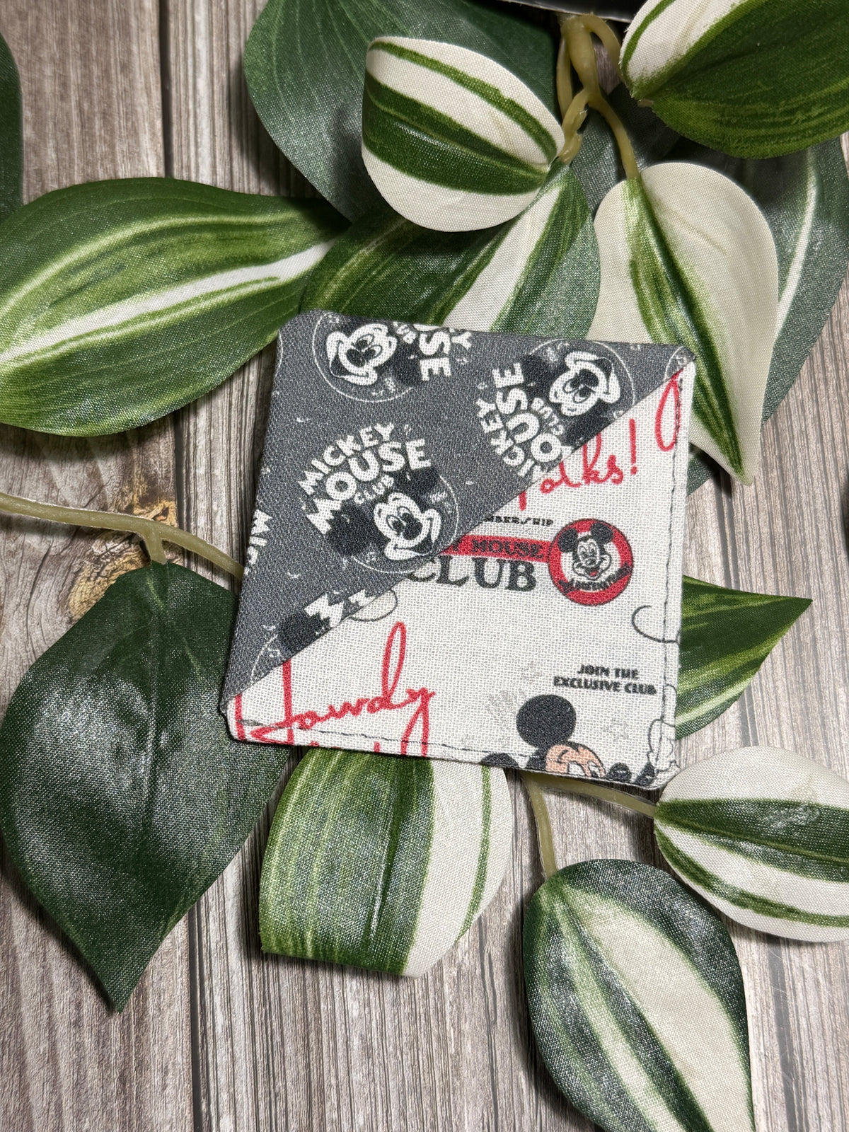 A square fabric book mark with a Mickey Mouse Club graphic print, styled on a rustic wood background with green and white variegated plant leaves.